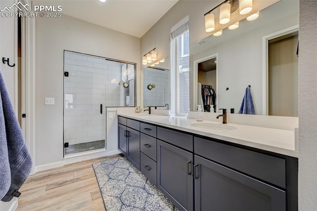 bathroom featuring wood finished floors, a shower stall, double vanity, a walk in closet, and baseboards