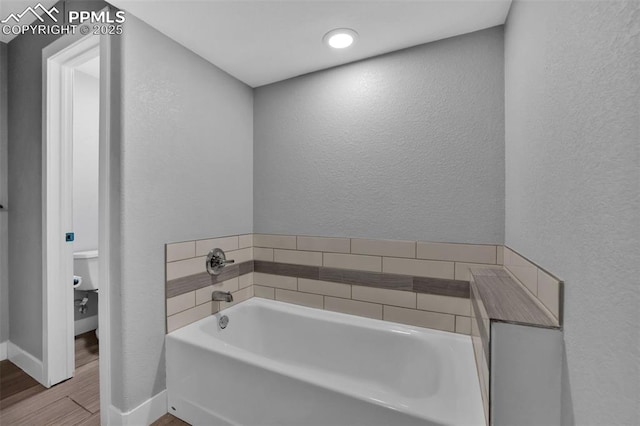 bathroom featuring wood finished floors, a bath, a textured wall, baseboards, and toilet