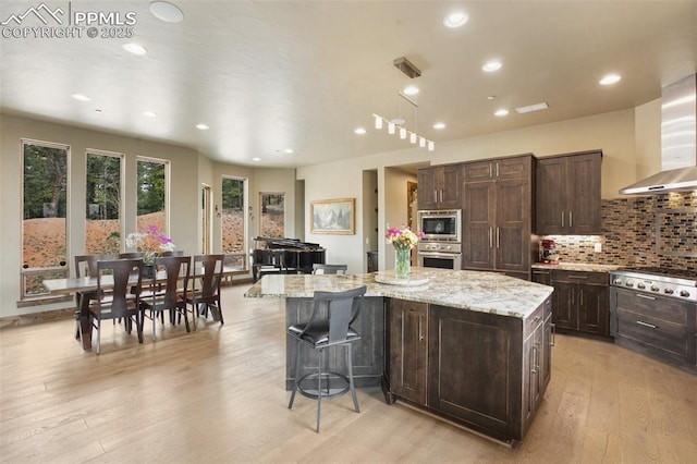 kitchen with appliances with stainless steel finishes, light wood-style floors, dark brown cabinets, a kitchen island, and light stone counters