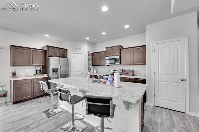 kitchen with appliances with stainless steel finishes, light wood finished floors, dark brown cabinets, a kitchen bar, and recessed lighting
