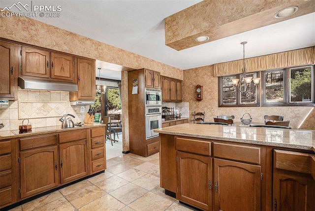 2518 Avondale Dr, Colorado Springs, CO 80917 - kitchen with a chandelier, brown cabinets, backsplash, under cabinet range hood, and hanging light fixtures