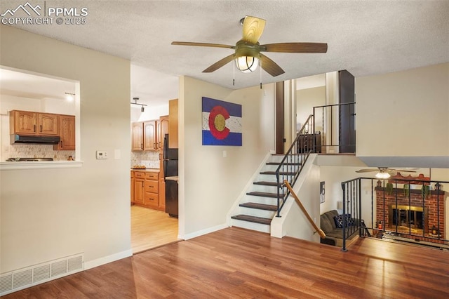 2417 Ranch Ln, Colorado Springs, CO 80918 - unfurnished living room featuring a ceiling fan, light wood-style floors, stairway, a textured ceiling, and a brick fireplace