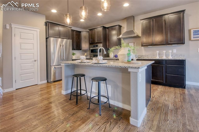 4228 Notch Trl, Colorado Springs, CO 80924 - kitchen featuring freestanding refrigerator, wall chimney exhaust hood, dark brown cabinetry, a center island with sink, and light wood finished floors