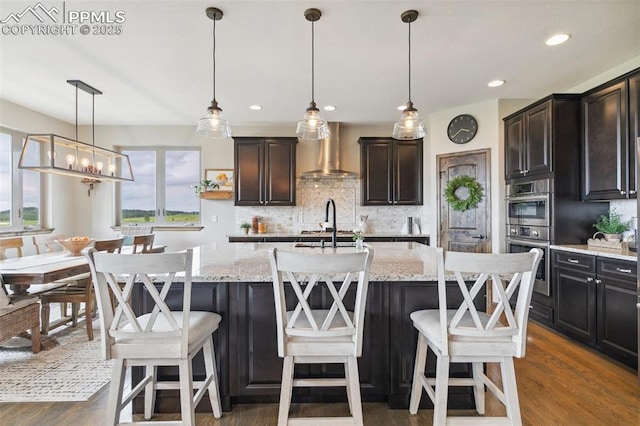 4808 Silver Nell Dr, Colorado Springs, CO 80908 - kitchen featuring wall chimney exhaust hood, stainless steel double oven, backsplash, dark wood finished floors, and a kitchen island with sink