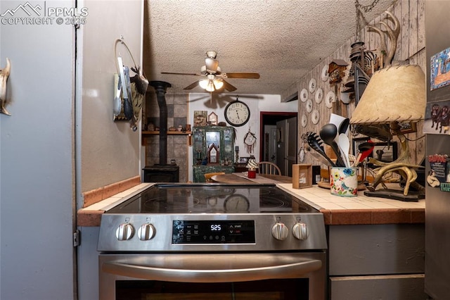 12185 Black Forest Rd, Colorado Springs, CO 80908 - kitchen featuring stainless steel electric range, tile counters, a textured ceiling, ceiling fan, and a wood stove