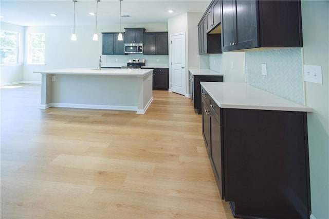 kitchen featuring a center island with sink, decorative backsplash, light wood-type flooring, appliances with stainless steel finishes, and decorative light fixtures