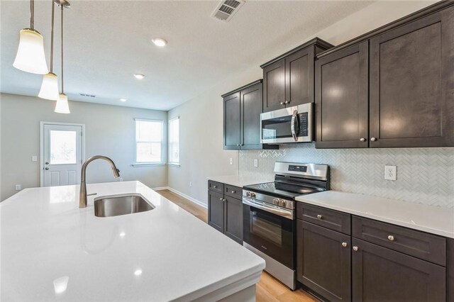 kitchen with sink, stainless steel appliances, light hardwood / wood-style floors, decorative light fixtures, and a center island with sink