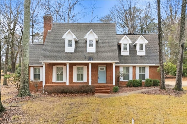 cape cod home with a chimney, roof with shingles, a porch, brick siding, and a front yard