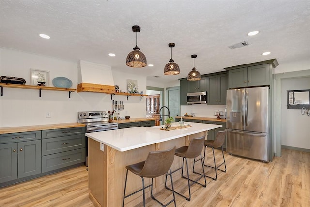 kitchen with light wood finished floors, custom exhaust hood, stainless steel appliances, open shelves, and visible vents