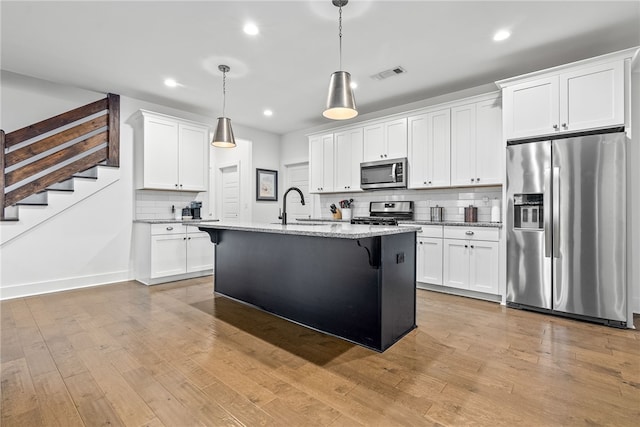 kitchen featuring appliances with stainless steel finishes, a kitchen island with sink, hanging light fixtures, light stone counters, and white cabinets