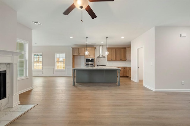 kitchen with appliances with stainless steel finishes, a fireplace, an island with sink, hanging light fixtures, and wall chimney range hood