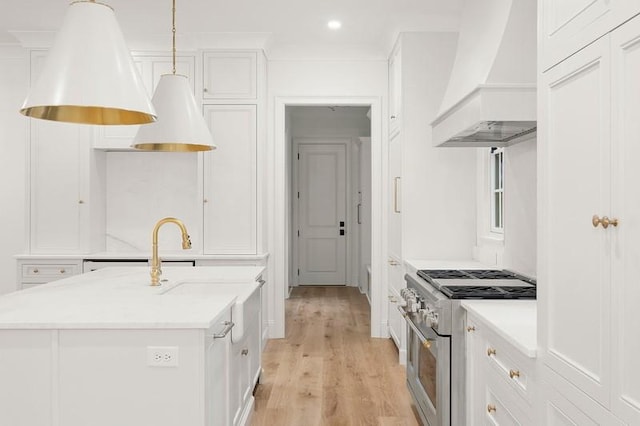 kitchen with white cabinets, stainless steel stove, custom range hood, hanging light fixtures, and a kitchen island with sink