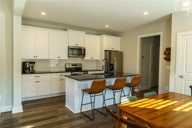 kitchen with a kitchen island with sink, appliances with stainless steel finishes, white cabinetry, dark countertops, and tasteful backsplash