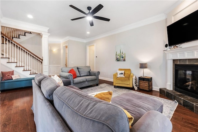 living area with a tiled fireplace, a ceiling fan, stairs, and wood finished floors