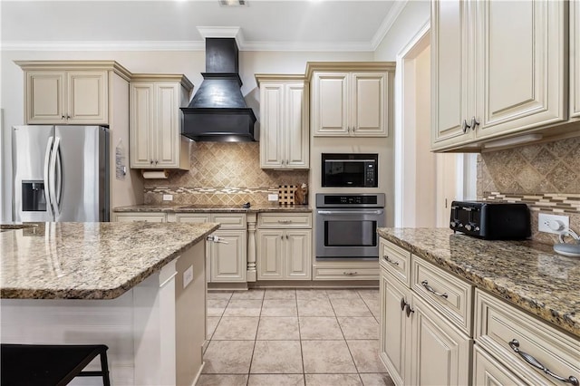 kitchen featuring light stone countertops, stainless steel appliances, decorative backsplash, cream cabinetry, and custom exhaust hood