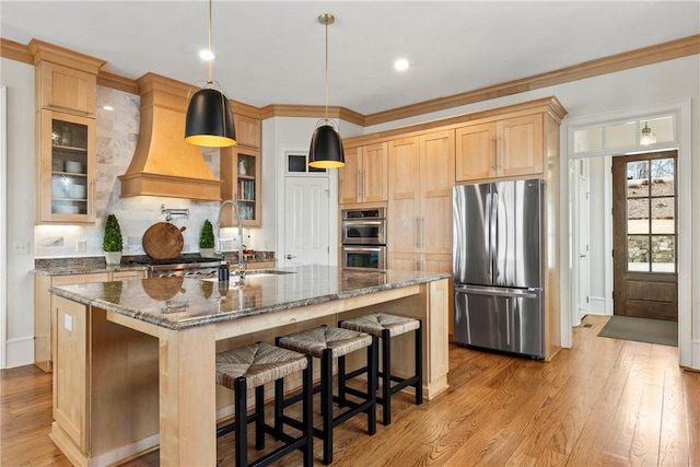 kitchen with appliances with stainless steel finishes, decorative light fixtures, an island with sink, dark stone countertops, and custom range hood