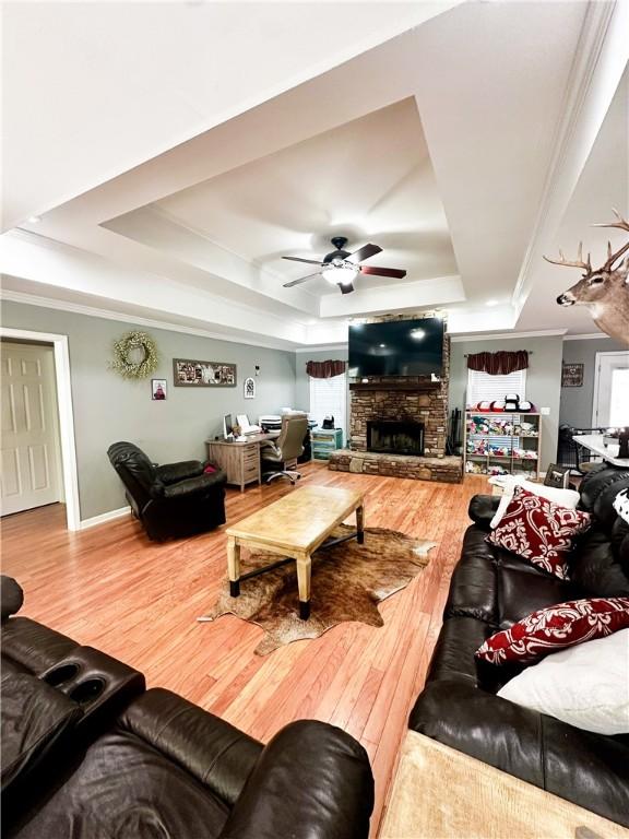 living room featuring hardwood / wood-style flooring, a raised ceiling, ornamental molding, ceiling fan, and a stone fireplace