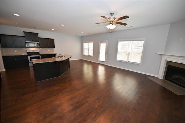 kitchen featuring a tile fireplace, a center island with sink, ceiling fan, light stone countertops, and stainless steel appliances