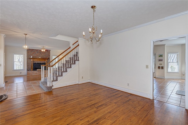 unfurnished living room with wood-type flooring, a textured ceiling, ornamental molding, a fireplace, and ceiling fan with notable chandelier