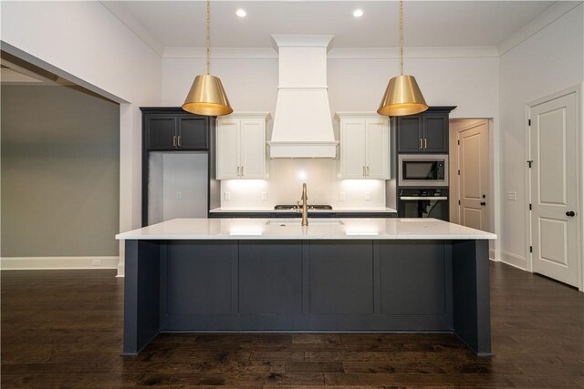 kitchen featuring stainless steel microwave, oven, an island with sink, decorative light fixtures, and custom range hood