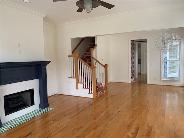 unfurnished living room with ceiling fan, light wood finished floors, a fireplace with flush hearth, stairs, and crown molding