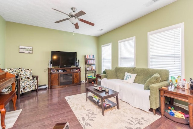 living area featuring visible vents, dark wood-type flooring, a glass covered fireplace, and a ceiling fan