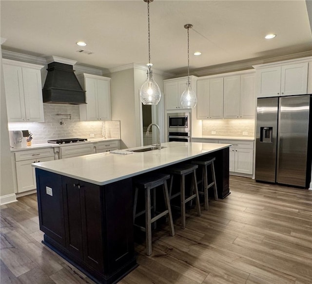 kitchen featuring a large island with sink, stainless steel appliances, white cabinetry, and premium range hood