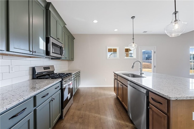 kitchen featuring light stone counters, stainless steel appliances, a kitchen island with sink, sink, and hanging light fixtures