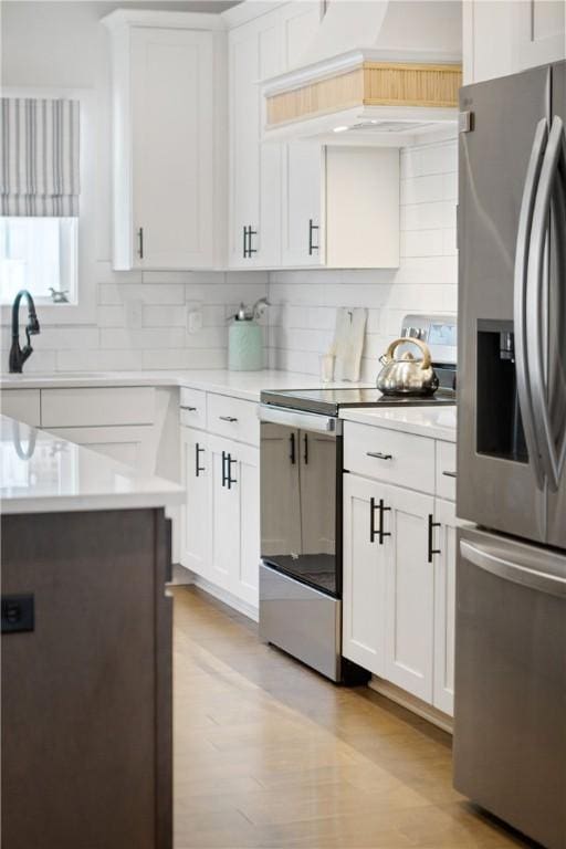 kitchen featuring light hardwood / wood-style flooring, premium range hood, appliances with stainless steel finishes, white cabinets, and decorative backsplash