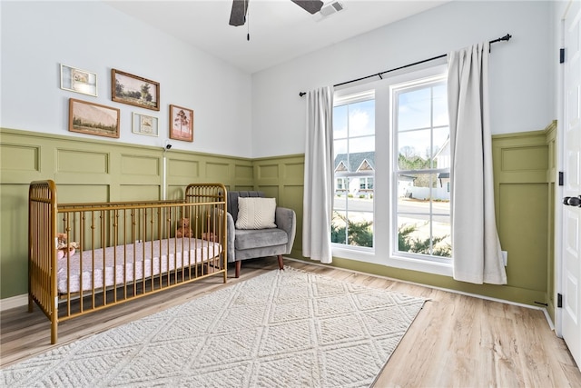 bedroom featuring wood finished floors, visible vents, a nursery area, wainscoting, and a decorative wall