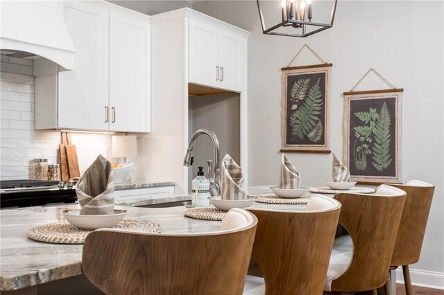 kitchen with decorative backsplash, light stone counters, custom exhaust hood, white cabinetry, and hanging light fixtures