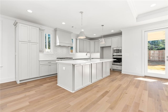 kitchen featuring light wood-type flooring, custom exhaust hood, stainless steel appliances, a kitchen island with sink, and pendant lighting