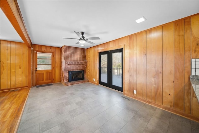 unfurnished living room featuring wood walls, a fireplace, visible vents, a ceiling fan, and french doors