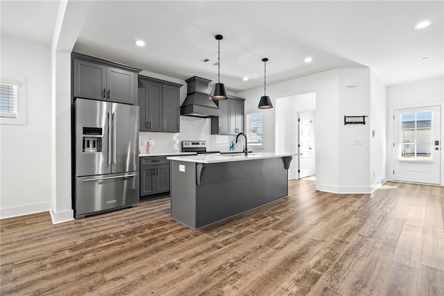 kitchen featuring appliances with stainless steel finishes, decorative light fixtures, an island with sink, sink, and custom exhaust hood