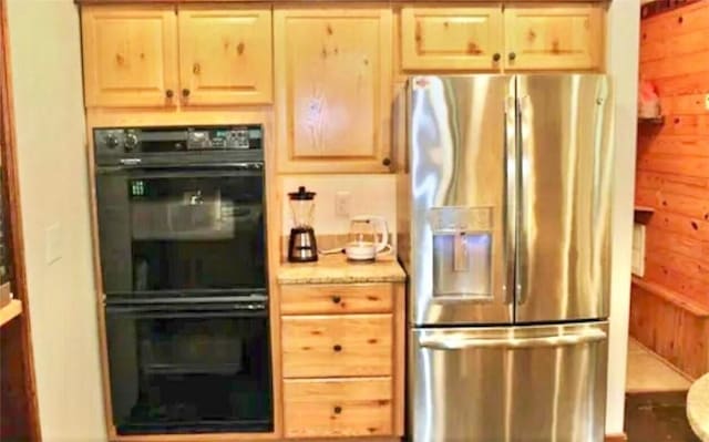 kitchen featuring light brown cabinetry, stainless steel fridge, black double oven, and wood walls