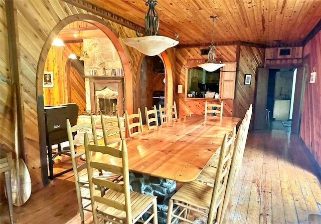 dining space with a stone fireplace, wood-type flooring, wooden ceiling, and wooden walls