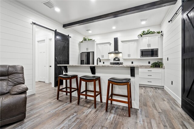 kitchen featuring a breakfast bar, wall chimney exhaust hood, a barn door, a center island with sink, and appliances with stainless steel finishes