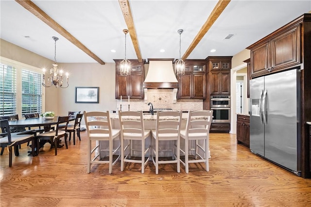 kitchen with appliances with stainless steel finishes, decorative light fixtures, a kitchen island with sink, and custom range hood