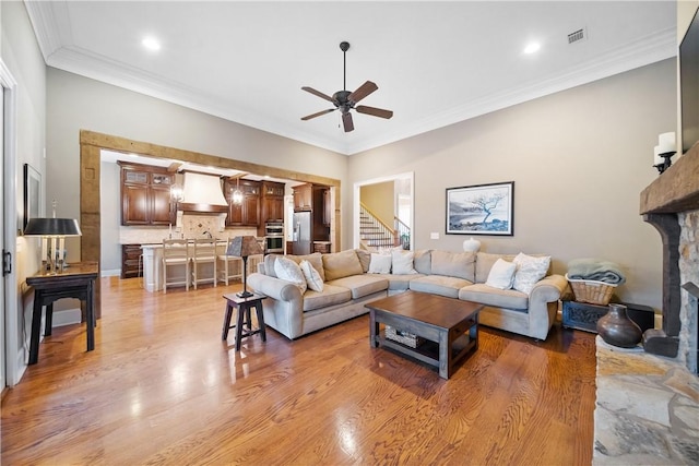 living room featuring a stone fireplace, sink, ornamental molding, ceiling fan, and light hardwood / wood-style floors