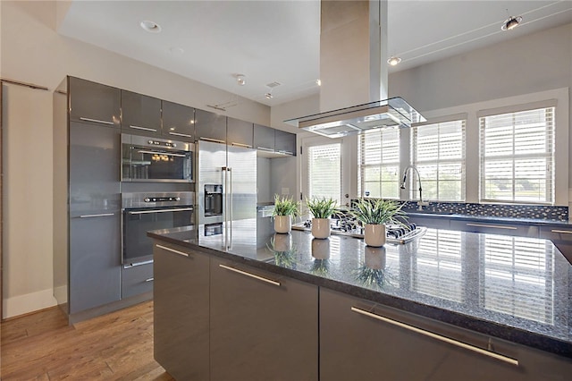 kitchen with island exhaust hood, light wood-style flooring, dark stone countertops, modern cabinets, and high end refrigerator
