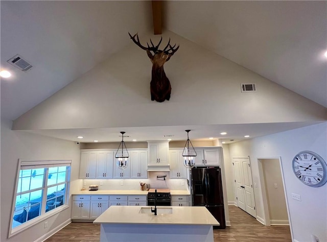 kitchen featuring an island with sink, white cabinets, hanging light fixtures, black fridge, and beam ceiling