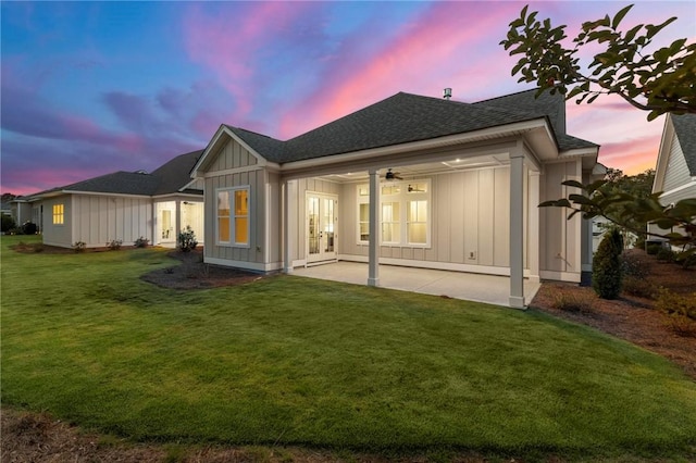 back of property at dusk featuring a shingled roof, board and batten siding, french doors, a ceiling fan, and a lawn