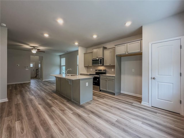 kitchen featuring a sink, light countertops, appliances with stainless steel finishes, gray cabinets, and a center island with sink