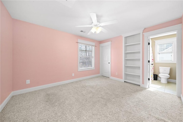 unfurnished bedroom featuring ceiling fan, light colored carpet, ensuite bath, and multiple windows