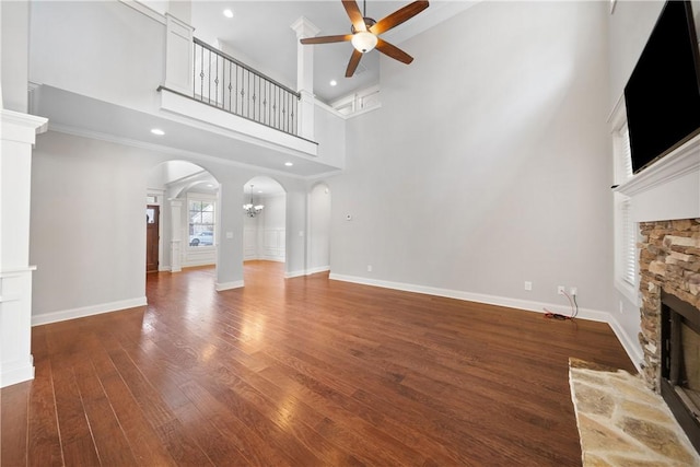 unfurnished living room featuring dark wood-type flooring, a stone fireplace, crown molding, ceiling fan with notable chandelier, and a high ceiling