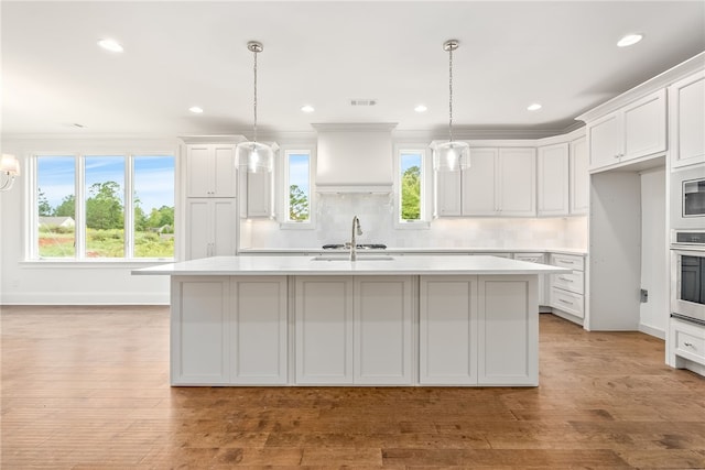 kitchen featuring pendant lighting, stainless steel oven, a kitchen island with sink, light hardwood / wood-style floors, and white cabinetry