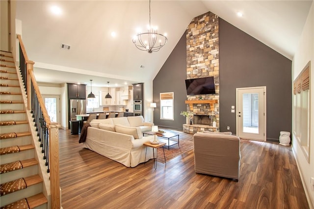 living room featuring a notable chandelier, high vaulted ceiling, dark wood-type flooring, stairs, and a stone fireplace