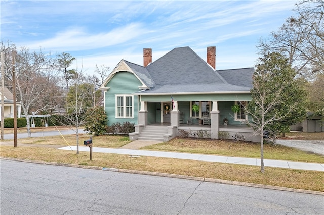 view of front facade with a front yard, covered porch, roof with shingles, and a chimney