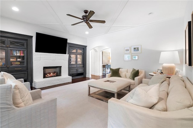 living area with a warm lit fireplace, arched walkways, coffered ceiling, a ceiling fan, and wood finished floors