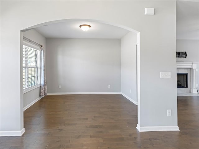 unfurnished room featuring arched walkways, a fireplace with flush hearth, baseboards, and wood finished floors